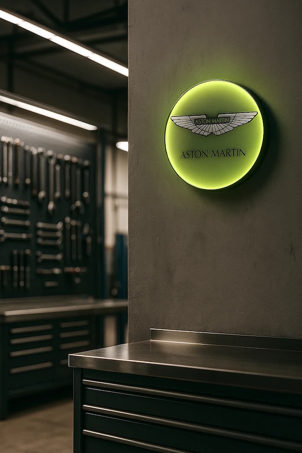 Aston Martin Logo Leuchte in grün mit LED Licht an einer grauen Wand in einer Werkstatt mit Ablageflächen und Werkzeug aus Metall
